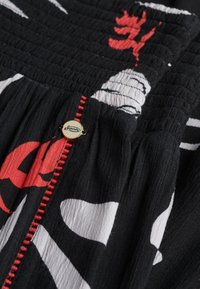 Sort stof med røde og hvide abstrakte mønstre, med rød syning og en rund cremefarvet knap med "Superdry"-logo i sort skrift.