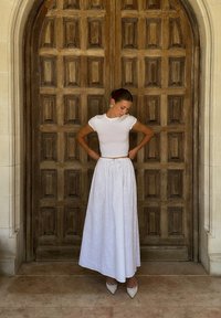 Weißes, kurzes T-Shirt kombiniert mit einem langen weißen Rock mit Durchbrüchen. Das Model trägt beigefarbene, spitze High Heels. Hintergrund aus Holzpanelen.