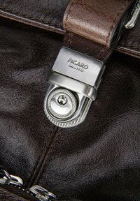 Détail d'un sac à main en cuir marron avec un fermoir en métal argenté présentant un motif circulaire texturé et un logo de marque gravé.