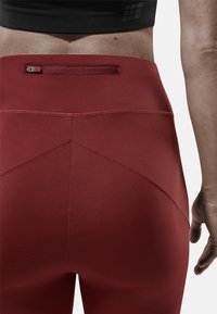 Leggings de sport rouges avec une texture lisse, dotés d'une poche zippée à l'arrière et de coutures décoratives pour un design ajusté.