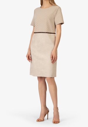 Femme portant une robe beige longueur aux genoux avec des manches courtes à motifs et des sandales à talons hauts couleur nude, debout sur un fond blanc.