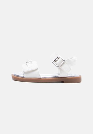Biały sandał z odkrytymi palcami, z dwoma regulowanymi srebrnymi klamrami i jasnobrązową płaską podeszwą, pokazany z boku na jednolitym tle.