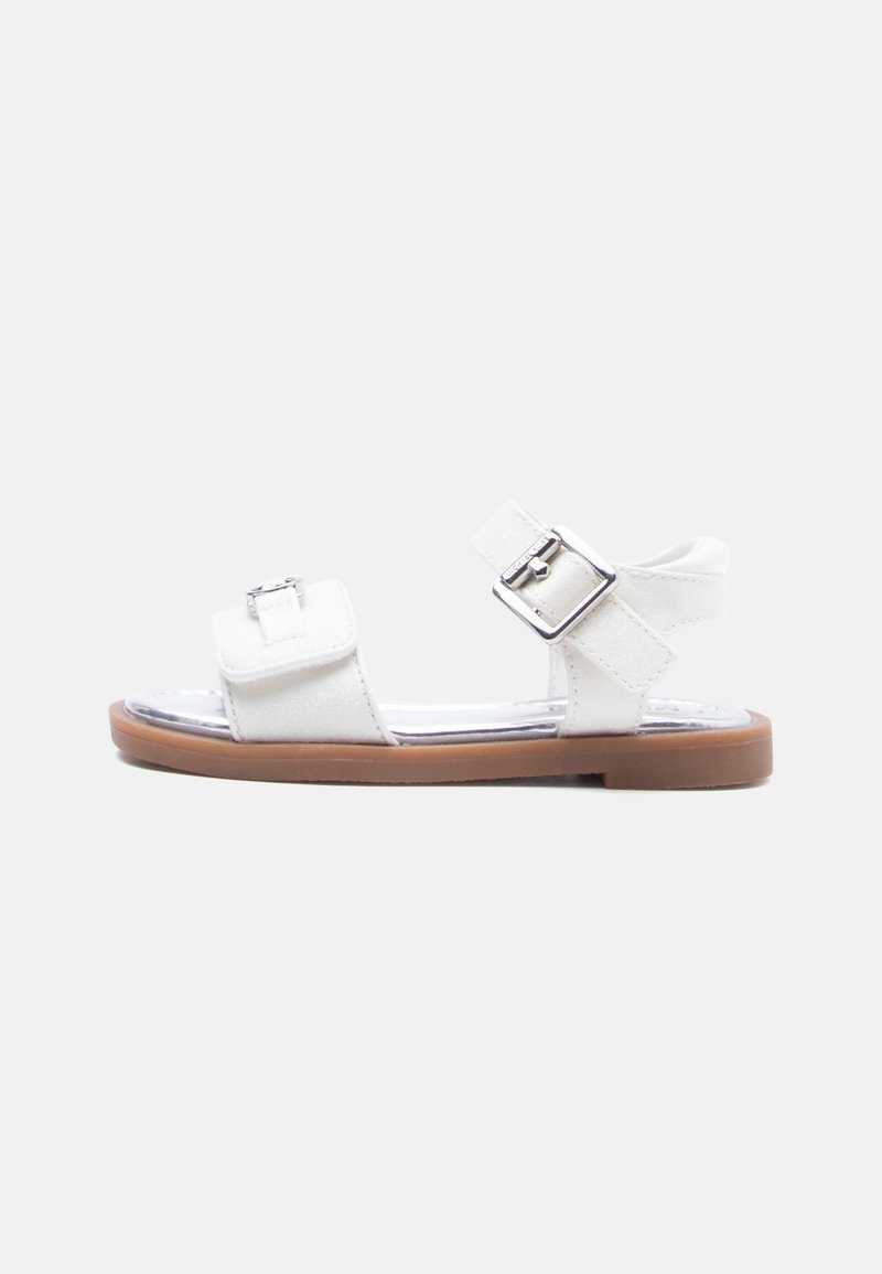 Λευκό σανδάλι ανοιχτού δαχτύλου με δύο ρυθμιζόμενες ασημένιες αγκράφες και καφέ επίπεδη σόλα, εμφανιζόμενο από το πλάι σε απλό φόντο.
