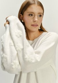 Witte gestructureerde trui met bloemenaccenten, met ribgebreide manchetten en een ontspannen pasvorm. De stof lijkt zacht en comfortabel.