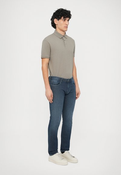 Šedé polokošeľa s krátkym rukávom a textúrovaným vzorom, v kombinácii s tmavomodrými slim-fit džínsami a bielymi teniskami. Mužská postava stojaca v profile.