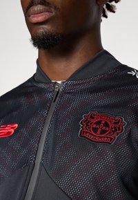 Svart träningsjacka med röda accenter och en strukturerad yta. Har en hel dragkedja, ståkrage och broderad Bayer Leverkusen-logotyp.