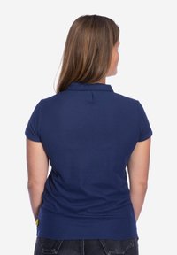 Marineblauwe polo gemaakt van katoen. Kenmerken zijn een kraag, korte mouwen en een gele accenttag aan de zoom. Achteraanzicht weergegeven.