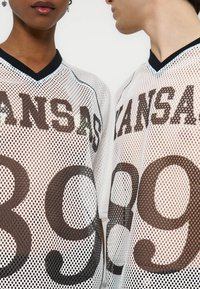 Δύο άτομα φορούν λευκά αθλητικά φανελάκια από δίχτυ με μαύρο περίγραμμα, που αναγράφουν «ZANSAB» και αριθμό 89 μεγάλο στην μπροστινή πλευρά.