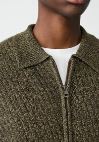 Detailný záber na osobu, ktorá má na sebe olivovo zelený textúrovaný pletený sveter na zips cez bielu košeľu, pričom sú viditeľné detaily goliera a zipsu.