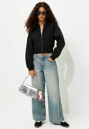 Joven con cabello rizado lleva chaqueta negra con cremallera, vaqueros de pierna ancha azul claro, zapatillas negras, gafas de sol oscuras y sostiene un bolso plateado.