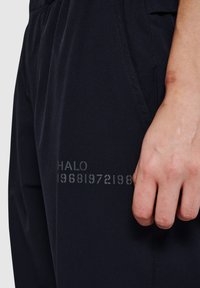 Schwarze Hose mit glatter Textur, die bedruckten Text "HALO 196819721988" an der Seite aufweist und damit Gestaltung und Markenmerkmale hervorhebt.