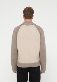 Homem de costas, vestindo um suéter de malha bicolor em bege e taupe, com colarinho, punhos e barra canelados, combinado com calças pretas.