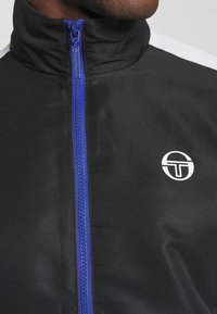 Melnā zipa jaka ar augstu apkakli, kurai ir zils zipa rāvējslēdzējs un balts logotips kreisajā pusē. Strukturēta auduma, ar kontrastējošām plecu svītrām.