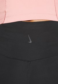 Czarne legginsy sportowe o gładkiej strukturze, z subtelnym szarym logo Nike w pobliżu pasa; noszone z jasnoróżową dopasowaną bluzką.
