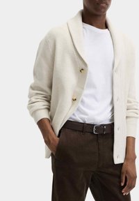 Cremfarbener Strickcardigan mit einem Schalkragen und goldenen Knöpfen. Kombiniert mit einem weißen T-Shirt und braunen Cordhosen. Strukturierter Stoff.