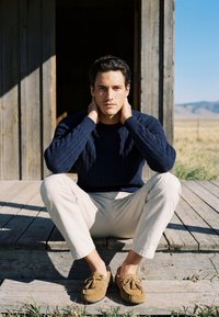 Homme en pull bleu marine et pantalon beige assis sur des marches de porche en bois, mains sur la nuque, paysage en arrière-plan.