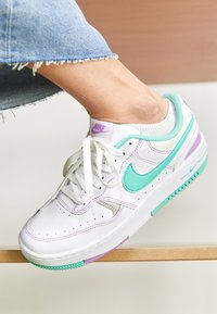 White Nike sneakers with teal and purple accents, featuring a textured upper, round toe, and flat laces. Perforated detailing for breathability.