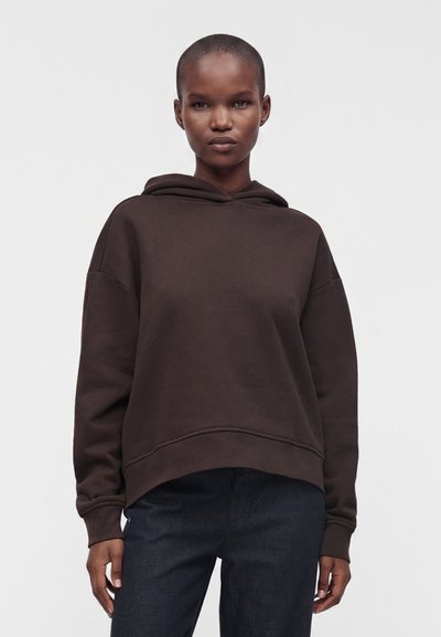 Personne debout face à l'avant, portant un sweat à capuche ample marron foncé et un pantalon foncé, avec une expression neutre et les cheveux rasés. Fond uni.