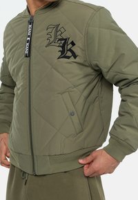 Giacca bomber trapuntata verde oliva con loghi neri ricamati, chiusura frontale con zip e polsini e orlo a coste. Presenta tasche laterali.