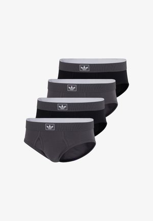 Młody mężczyzna ze średniej długości dredami, ubrany w ciemnoszare slipy, stojący boso na białym tle