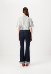 Chemise grise à manches courtes associée à un jean flare bleu foncé avec ourlets roulés et poches arrière. Chaussures à talons hauts rouges visibles.