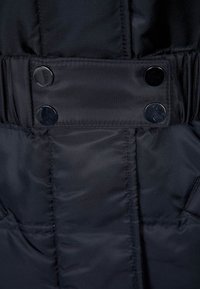 Svart vadderad jacka med en glänsande panel som har två stora svarta tryckknappar och strukturerad tyg, som framhäver sömnads- och sömspecifikationer.