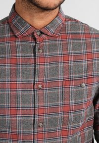 Camisa de franela a cuadros en rojo y gris con cuello abotonado y botones grises, con una textura suave y líneas horizontales y verticales en contraste.