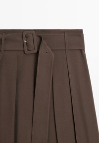 Hnědá plisovaná sukně se širokým látkovým páskem a obdélníkovou sponou, stylizovaná s poutky na pásek na bílém pozadí.