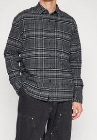 Camisa de cuadros gris y negro, confeccionada en tejido, con diseño de botones, mangas largas y un bolsillo en el pecho. Acabado texturizado.