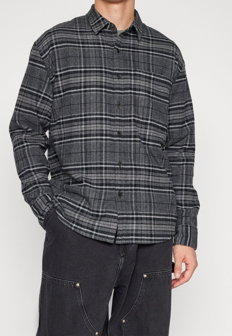 Camisa de cuadros gris y negro, confeccionada en tejido, con diseño de botones, mangas largas y un bolsillo en el pecho. Acabado texturizado.