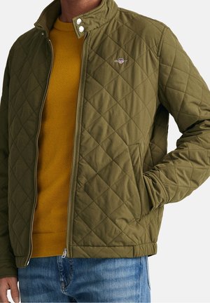 Mand iført olivengrøn quiltet jakke over sennepsweater og blå jeans, den ene hånd i jakkelommen.