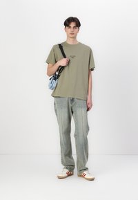 Olive green cotton t-shirt with printed logo, faded blue jeans, and cream sneakers with red stripes. Model holds a light blue crossbody bag.