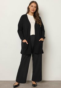 Schwarze, offene Strickjacke mit zwei Fronttaschen, langen Ärmeln und einem lockeren Schnitt, kombiniert mit schwarzen Hosen und einem hellen Oberteil.