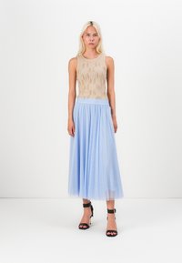 Beige strikket tanktopp med teksturert mønster, kombinert med en lyseblå plisserte skjørt. Svarte sandaler med stropper fullfører antrekket.