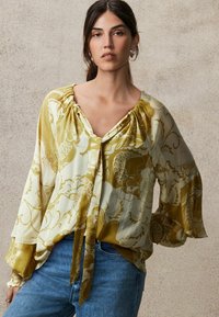 Bluză de culoare galben deschis cu modele florale aurii, cu mâneci bufante și decolteu în V. Prezintă un nod în față și un material texturat. Purătă cu blugi.