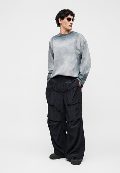 Νέος άνδρας που φορά γκρι μακρυμάνικο πουκάμισο, μαύρο oversized cargo παντελόνι, μαύρα αθλητικά παπούτσια και σκούρα γυαλιά ηλίου, στέκεται με τα χέρια στις τσέπες.