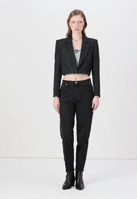 Blazer nero accollato con texture tonale e rever acuti, abbinato a jeans neri a vita alta e stivaletti, su uno sfondo neutro.