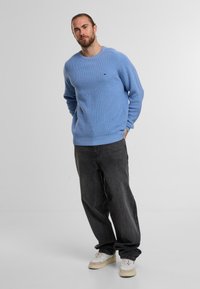 Mann mit Bart im hellblauen Pullover, schwarzen lockeren Jeans und weißen Sneakern, der mit hinter dem Rücken verschränkten Händen vor einem grauen Hintergrund steht.