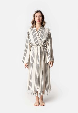 Femme debout pieds nus portant une longue robe blanche à rayures verticales noires avec des glands, nouée à la taille sur un fond uni.