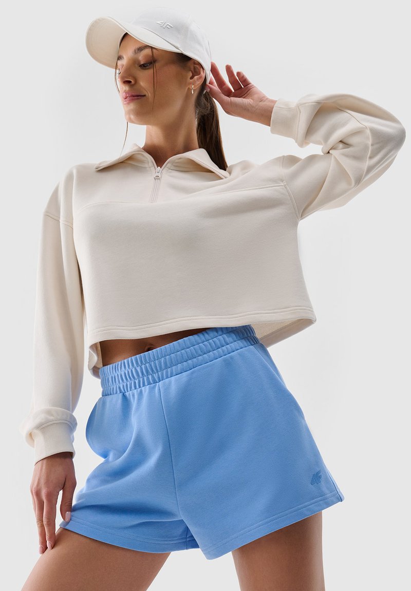 Femme portant un sweat-shirt court beige, un short bleu clair à taille élastique et une casquette blanche, posant avec un bras levé et la tête tournée.