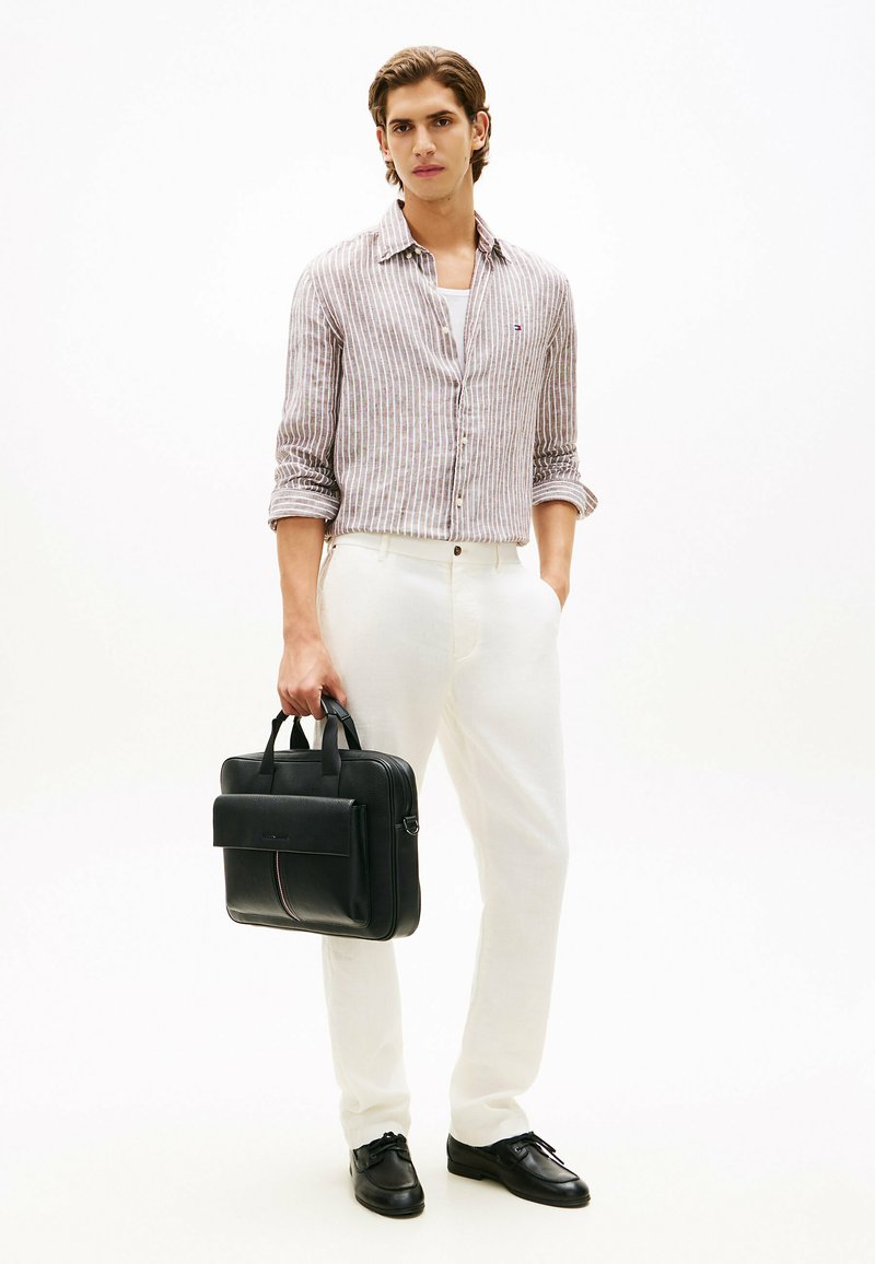 Jeune homme en pantalon blanc et chemise rayée tenant une mallette en cuir noir, debout devant un fond blanc uni.