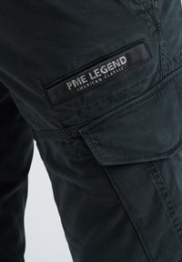 Dunkelgrüne Cargo-Hosen mit strukturiertem Finish, mehreren Taschen und einem Etikett mit der Aufschrift "PME LEGEND AMERICAN CLASSIC" an der Seite.