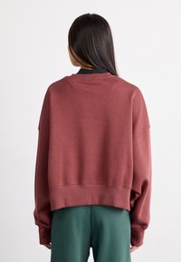 Osoba s dlouhými tmavými vlasy, oblečená v volném rustově červeném svetru a tmavě zelených kalhotách, zobrazená zezadu na jednobarevném bílém pozadí.