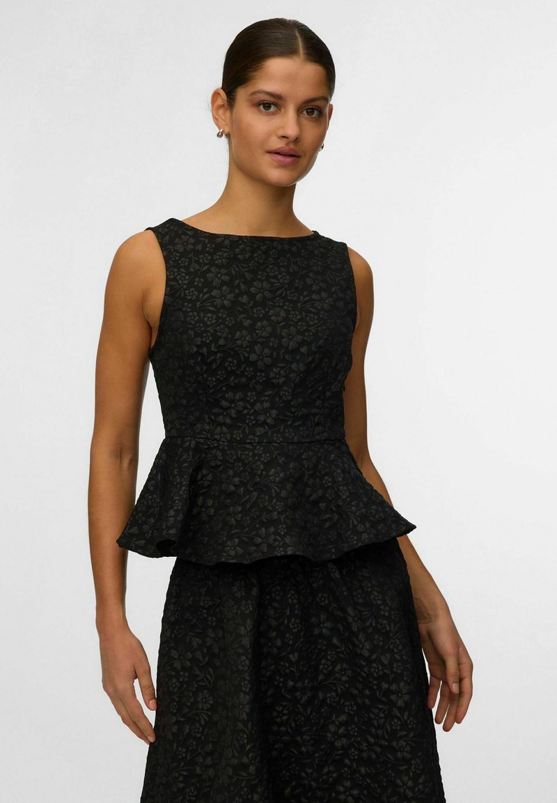 Femme portant un haut péplum sans manches à motif floral noir et une jupe assortie, debout devant un fond clair uni.