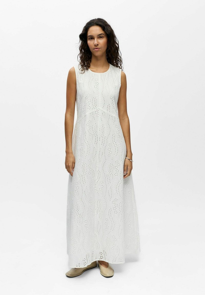 Femme aux cheveux bouclés foncés portant une robe maxi blanche sans manches avec des détails en broderie anglaise et des ballerines beiges, debout sur un fond blanc.