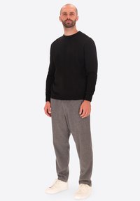 Uomo in piedi che indossa una camicia nera a maniche lunghe, pantaloni di lana grigi e sneakers bianche su uno sfondo leggero e uniforme.