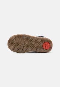 Suola di sneaker con una base in gomma marrone, battistrada texturizzato per la presa e un accento circolare rosso. Presenta scanalature pronunciate e il logo del marchio.