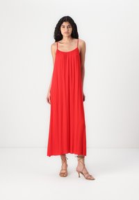 Robe maxi rouge avec des bretelles fines, un col froncé et un tissu fluide. Accompagnée de sandales à talons hauts beiges avec des lanières.