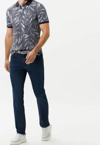 Man i mörkblå jeans, vita sneakers och en grå kortärmad piké med ett vitt bladmönster, står mot en vit bakgrund.