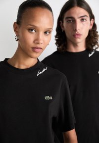 Sorte bomulls t-skjorter med rund hals; har brodert Lacoste-logo og hvit skrift på skulderen; glatt tekstur.